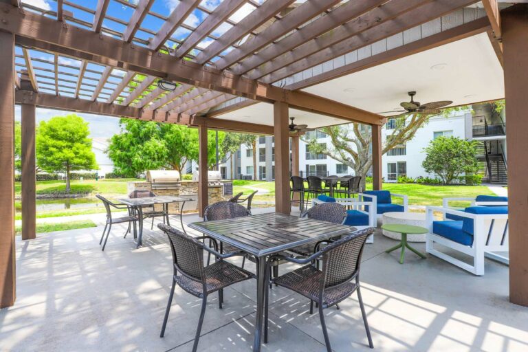 Outdoor gazebo and pergola with fans, tables and chairs and two grills next to a lake with apartment buildings in the background.