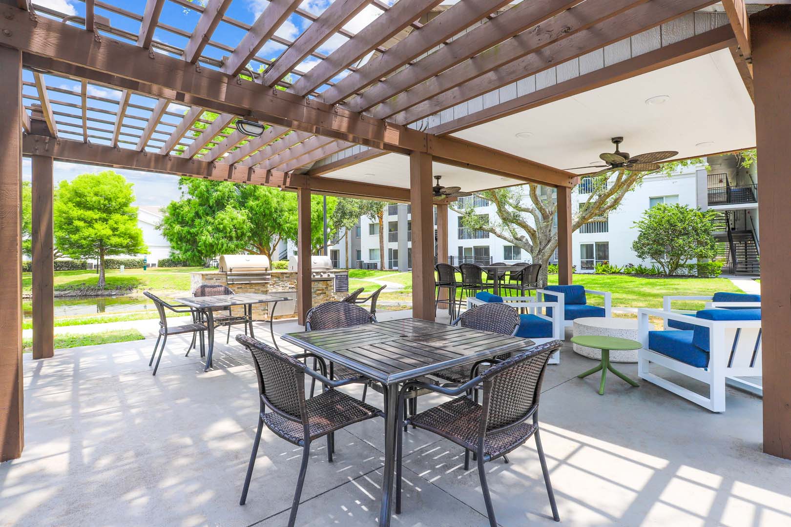 Outdoor gazebo and pergola with fans, tables and chairs and two grills next to a lake with apartment buildings in the background.