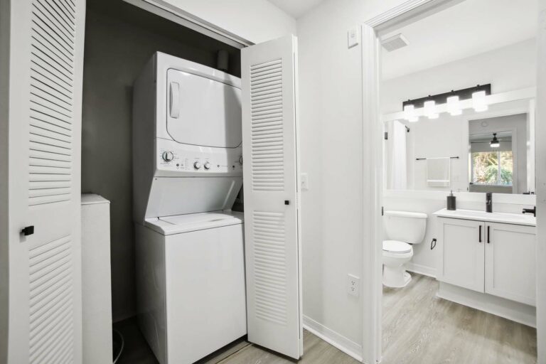 Laundry closet with stacked washer and dryer, next to bathroom with hardwood-style flooring, white shaker cabinets with white accents, vanity mirror with vanity lighting